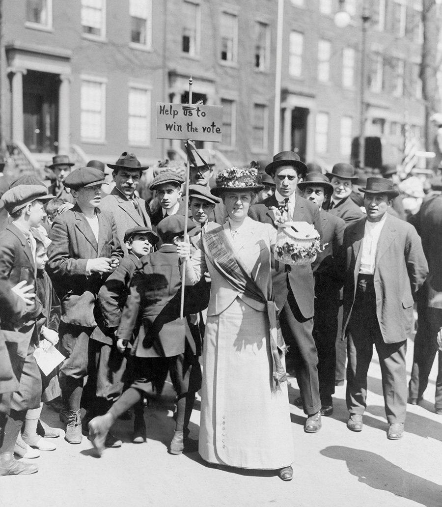 Women identified as Mrs. Suffern, is surrounded by a crowd of men and boys, while she holds a home-made banner in women suffragist parade ‘Help us to win the vote.’ 1914.